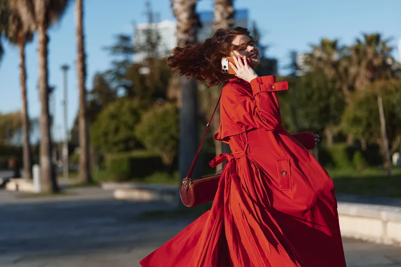 Woman in red trench coat walking outdoors while talking on smartphone, sunny day with palm trees in background. Casual urban lifestyle, communication concept.