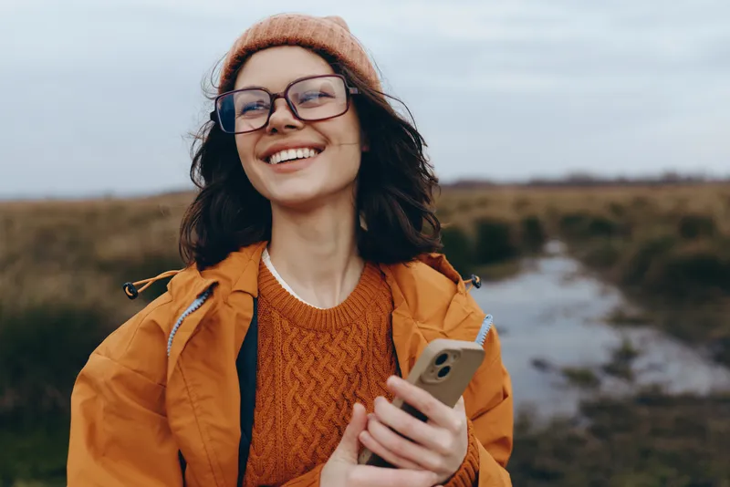 Woman outdoors smiling in sunlight, wearing hat and glasses, holding smartphone on a scenic landscape path, casual lifestyle travel moment, orange jacket and knit sweater