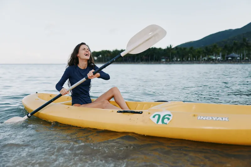 A joyful woman paddles a bright yellow kayak in calm coastal water, smiling as she enjoys a sunny outdoor moment.