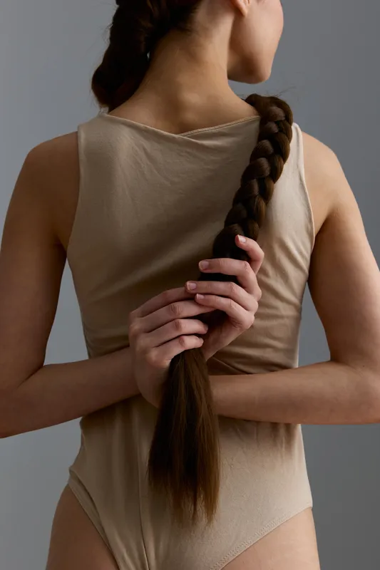 woman holding long braided hair behind back, beige clothing, smooth skin, minimalist gray background, natural hairstyle, soft lighting portrait in studio