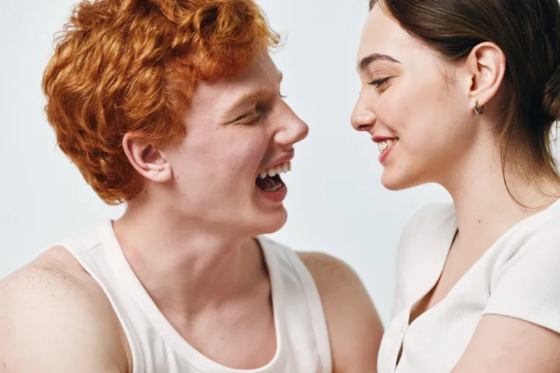 Young couple with red hair and dark hair facing each other smiling and laughing with happiness and joy on white background. Closeup of cheerful people in casual white clothes.