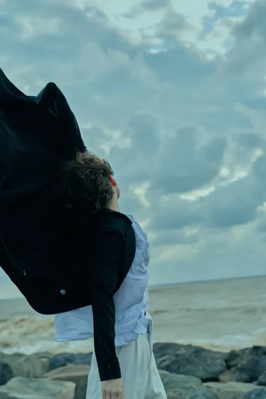 Silhouette of a figure near a windy coastal shore, lifting a dark jacket over the shoulder as dramatic clouds drift above rugged rocks and gray water, capturing motion and mood