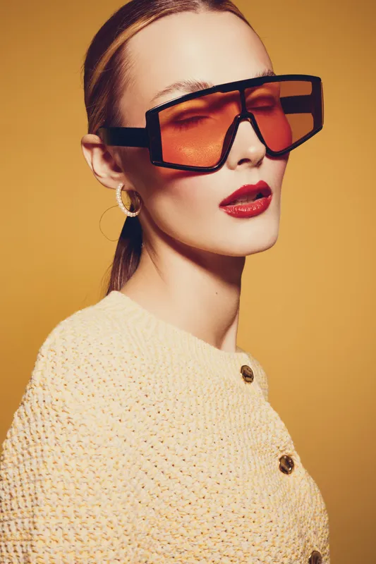 Stylish woman with oversized orange sunglasses and red lipstick poses against yellow background. She wears a textured cream sweater with buttons. Concept of fashion, beauty, and modern style.