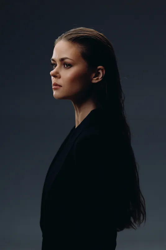 Profile of a serious woman with long hair wearing a black jacket against a dark background