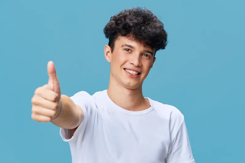 smiling man showing thumbs up gesture with friendly expression, casual white t-shirt, curly hair, blue background, positive mood, young adult