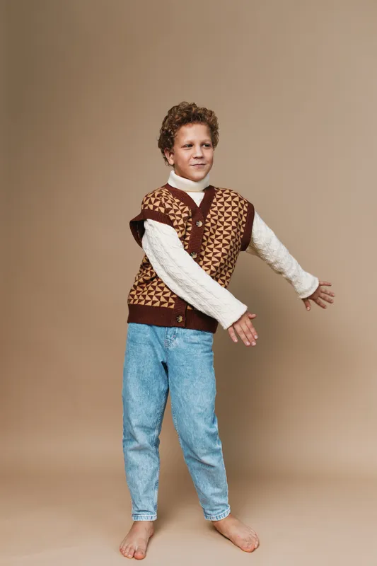 Playful child poses with arms outstretched, wearing a brown argyle knit vest over a white turtleneck and light jeans, set against a warm beige studio backdrop.