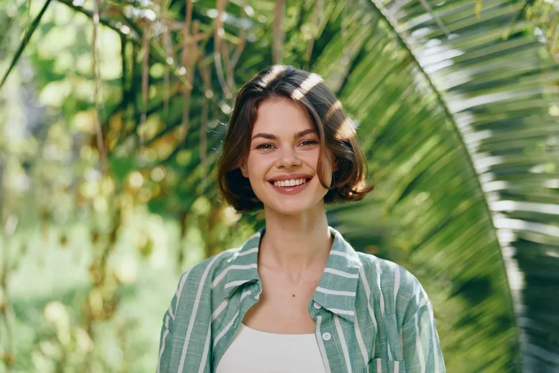Smiling young woman stands in a sunny tropical garden, wearing a striped green shirt over a white top, with lush foliage creating a fresh, casual mood.