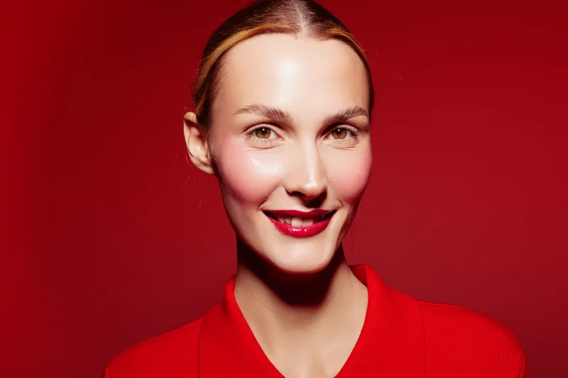 Smiling woman with red lipstick and blush wears red sweater against red background. Close-up portrait with soft lighting creates warm and vibrant mood. Concept of beauty and fashion.