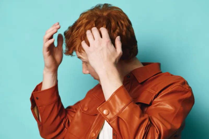 Redhead man in brown leather jacket adjusting hair with hands against blue background. Side profile, fashion style and casual clothing indoors with natural light.