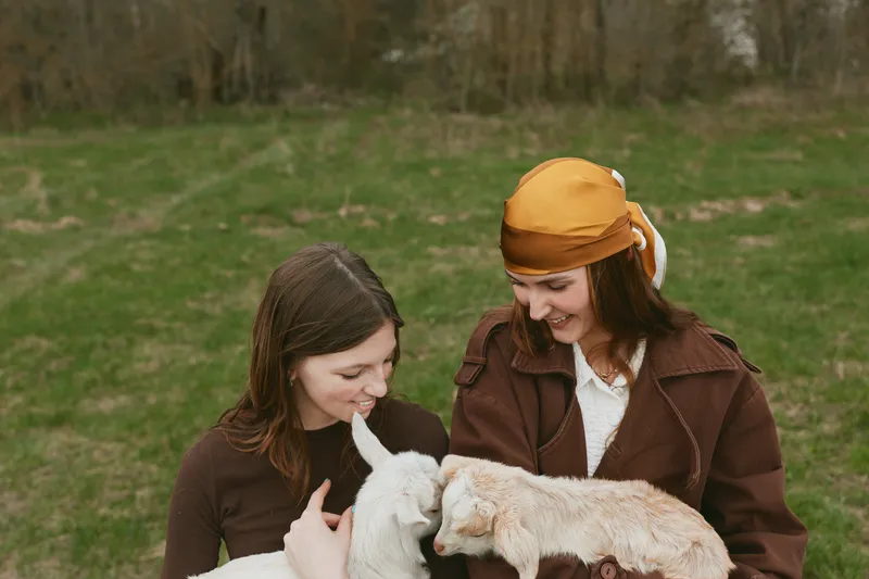 Lifestyle scene with two women outdoors, smiling as they interact with a pair of young goats, sunlit meadow, casual coats, gentle breeze, wholesome nature moment, candid connection, friendly