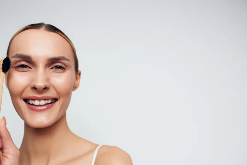 Smiling woman with makeup brush applying product to her cheek, close-up on a light neutral background. Concept of skincare, beauty, and self care with calm studio lighting