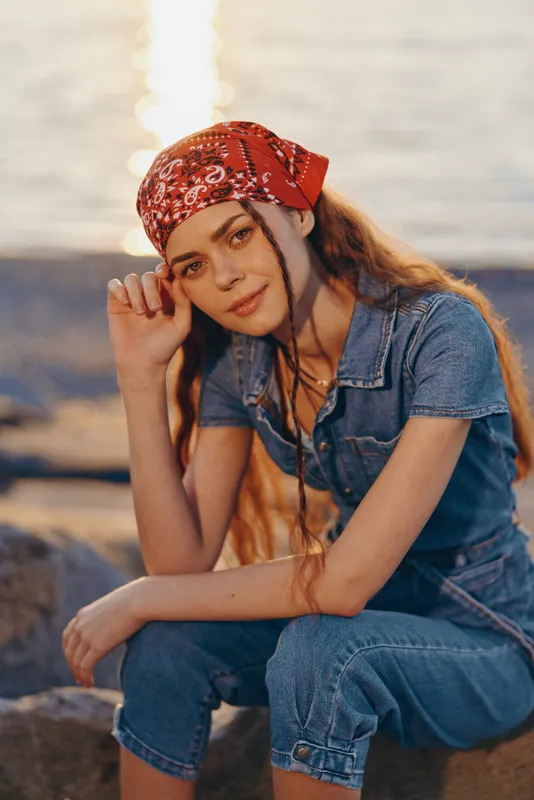 Young woman wearing red bandana and denim outfit sitting by the calm sea during sunset, relaxed and thoughtful in warm natural light.