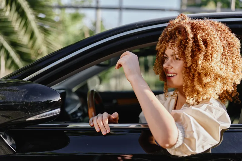 Smiling woman with curly hair enjoying driving a black car outdoors, happy expression, casual clothing, bright natural light, urban background.