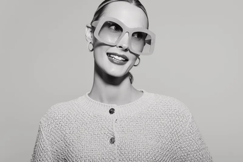 Smiling white woman with fair skin wearing oversized sunglasses and textured sweater poses against grey background. Concept of fashion, style, and confidence.
