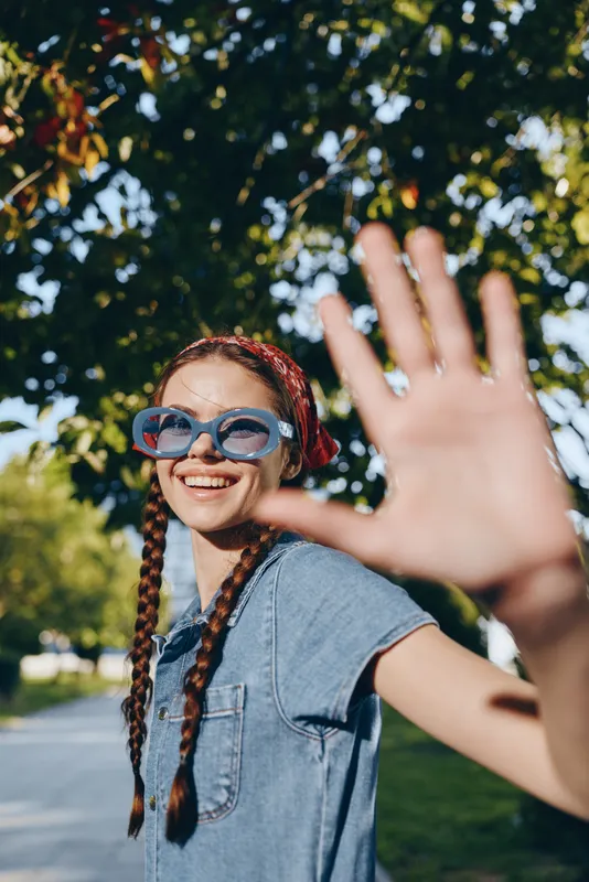 Lifestyle woman in modern boho-western denim street style wearing sunglasses with braided hair, smiling outdoors under natural sunlight, film color effect highlights warmth and joy.