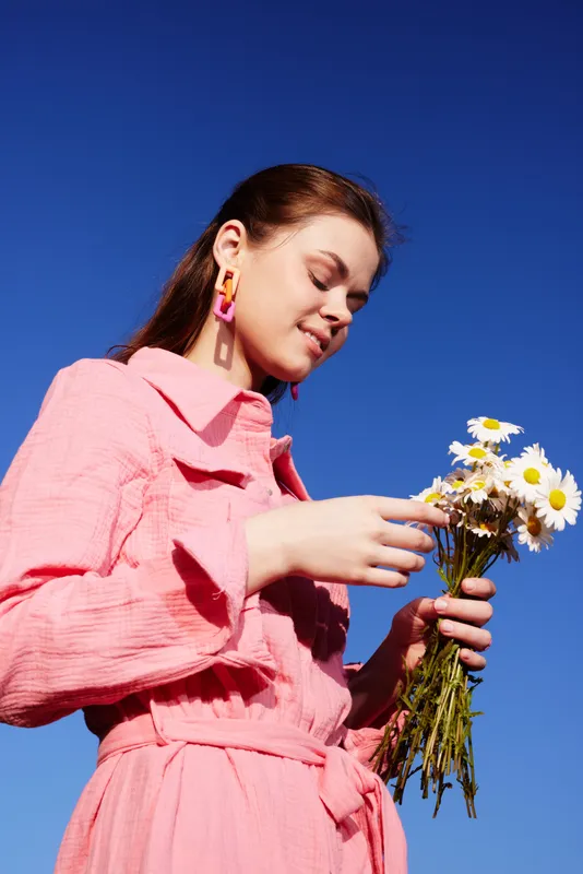 Young woman in pink dress holding daisies against a blue sky, embodying spring vibes and positivity with a joyful expression Bright colors enhance the cheerful mood