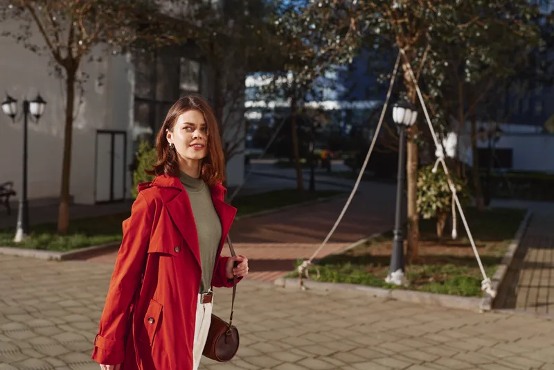 A young brunette woman in a red coat and green turtleneck walks along a sunlit paved urban park, smiling softly at the camera, with a crossbody bag swinging.