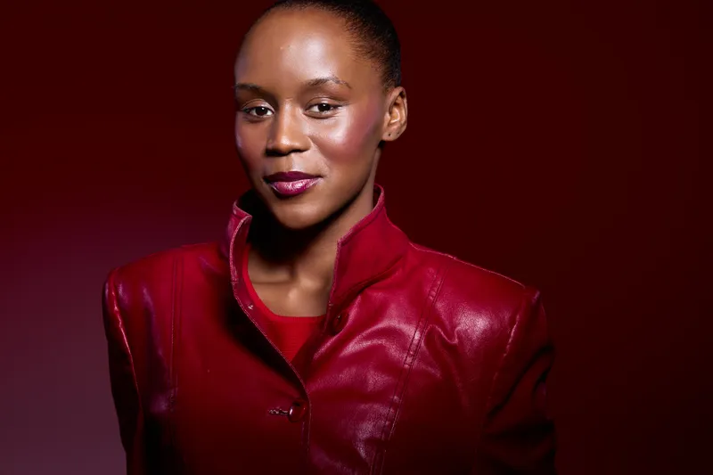 Confident African woman with dark skin wearing a red leather jacket poses against a red background. Studio portrait with strong lighting highlights elegance and fashion concept.