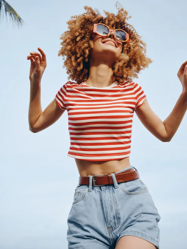 A carefree young woman in a red striped crop top and denim shorts dances beneath a clear blue sky, radiating joy, energy, and playful summer spirit.