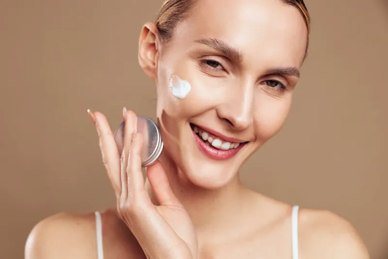 Woman skincare face cream beauty smile closeup, young woman applies moisturizer to cheek, gentle hand touch, studio lighting, clean skin glow, calm expression, neutral background