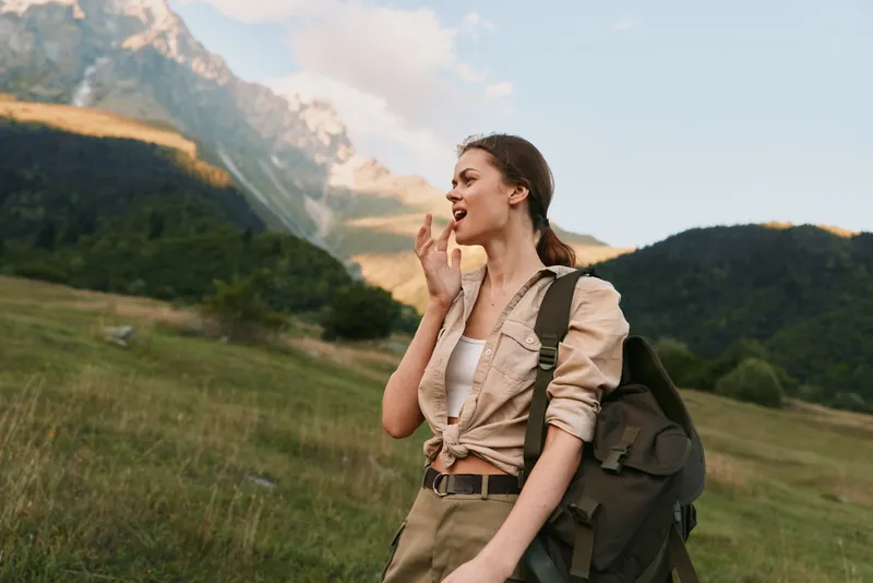 Young woman hiking outdoors with a backpack, mountains behind, enjoying nature and travel. Focus on lifestyle, exploration, wellness, and outdoor adventure.
