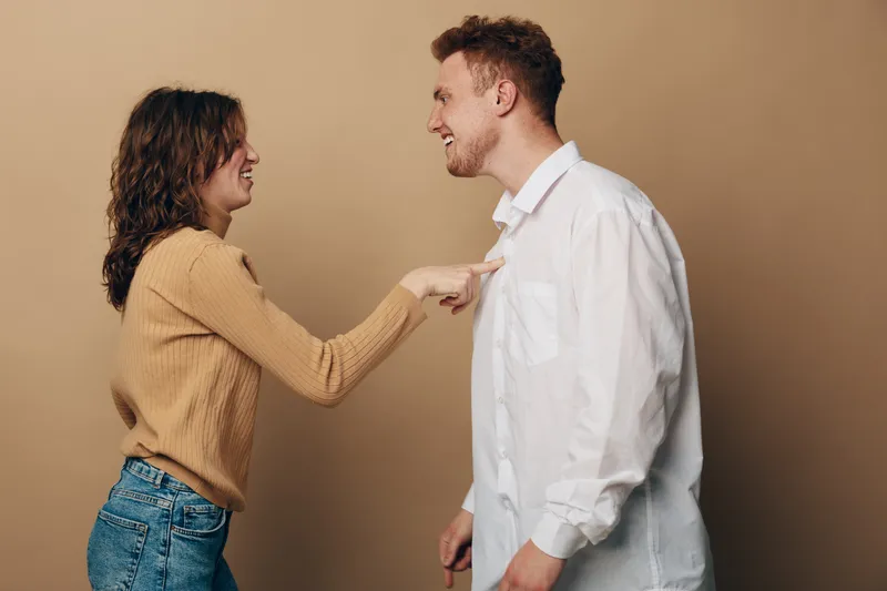 Smiling young woman in beige sweater playfully touches chest of laughing man in white shirt against beige background. Concept of happy couple, relationship, fun, and positive emotions.