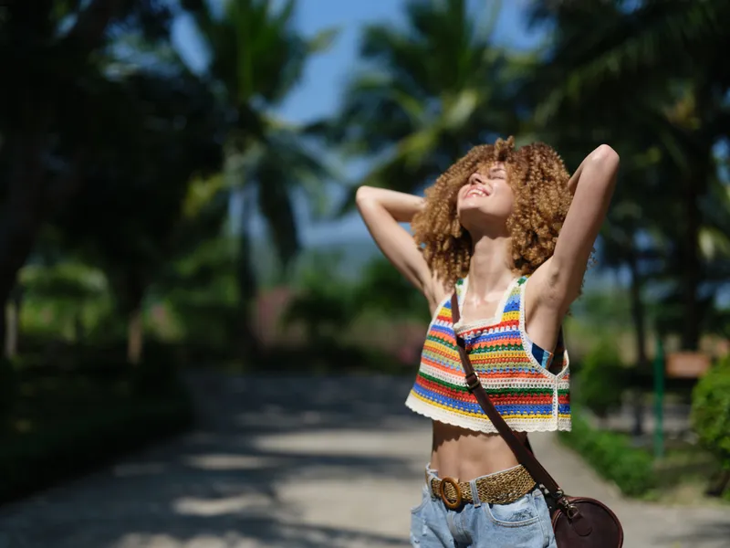 Smiling woman enjoying summer outdoors with relaxed pose, colorful striped top, and curly hair under bright sunlight near palm trees.