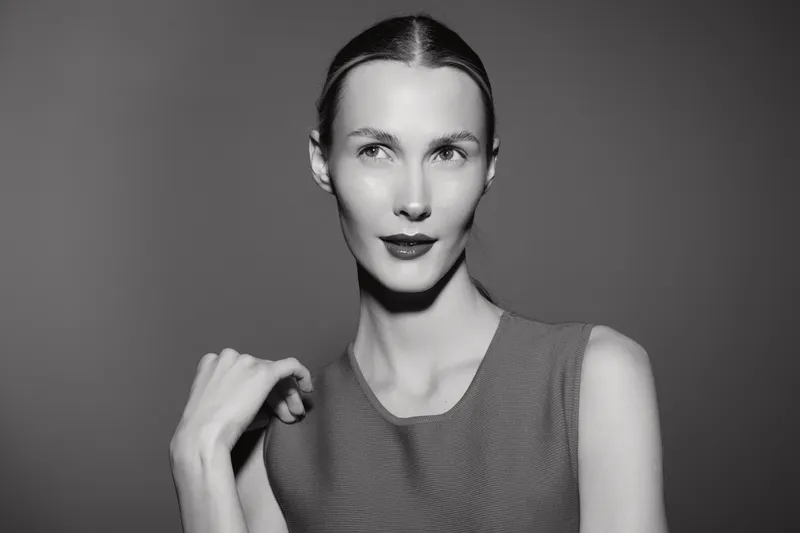 Portrait of a pale white woman with slicked hair wearing a sleeveless top posing against a plain background in black and white studio lighting.