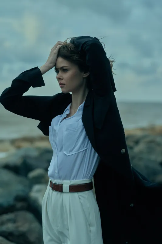 Woman in blazer and white pants stands on a windy shore, hair and coat swept by sea breeze, conveying determination, style and outdoor elegance, posed against rugged rocks and expansive sky,
