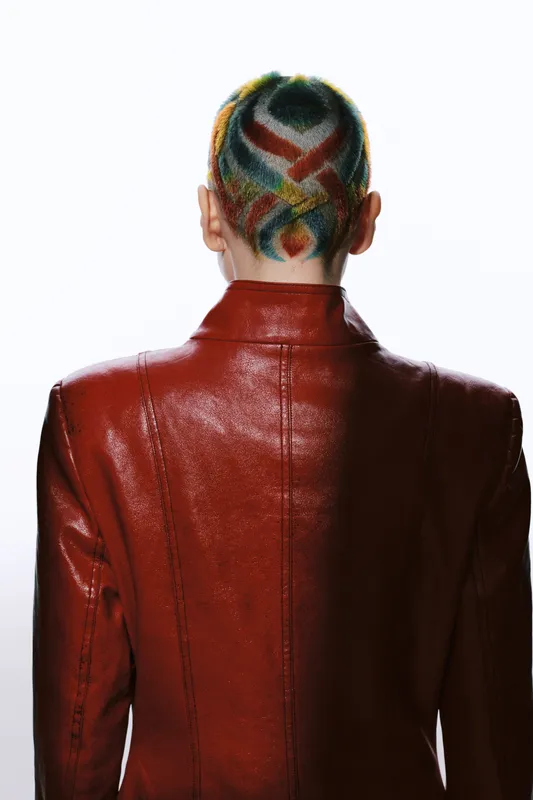 Woman with colorful shaved hair design wearing a shiny red leather jacket, viewed from the back against a white background, showcasing unique fashion and bold style.