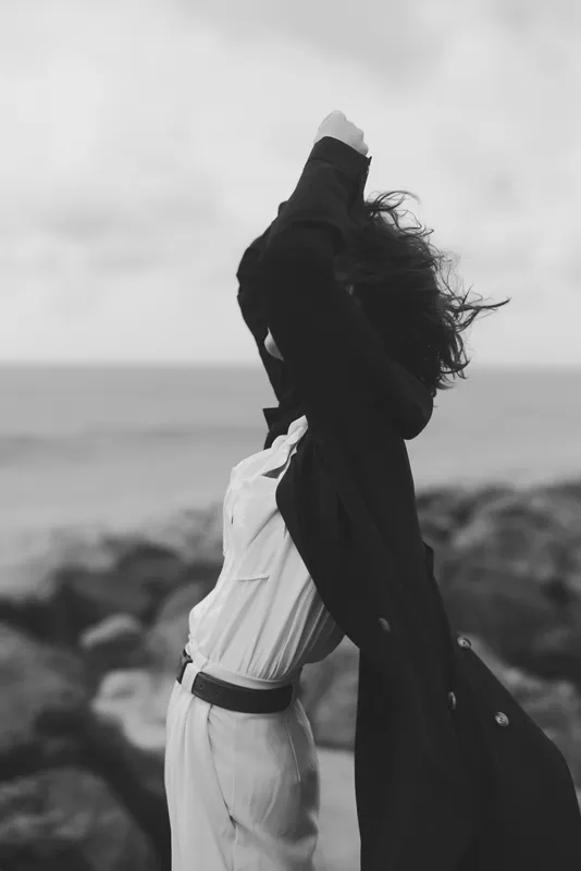 Freedom, wind, jacket, coast, ocean, movement, fashion, silhouette: a figure lifts a coat against a sea breeze on a rocky shore in monochrome