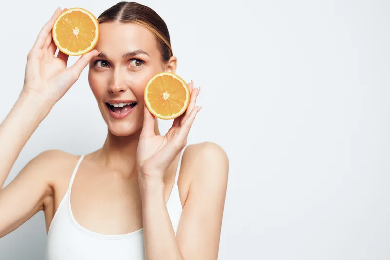 Young woman with long blonde hair holds two halves of an orange near her face and smiles playfully against a white background.