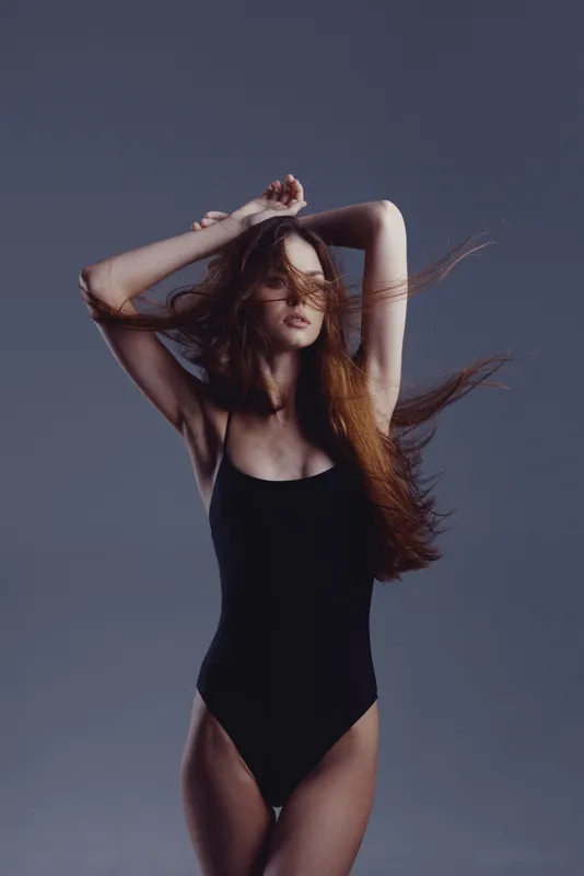 Young woman with long hair posing in black bodysuit against dark background, showing confident and elegant expression.