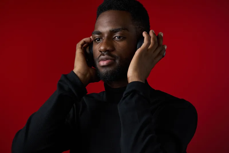 A focused young Black man in a black turtleneck, enjoying music with headphones against a vibrant red background, exuding calmness and concentration, ideal for themes of relaxation and mindfulness