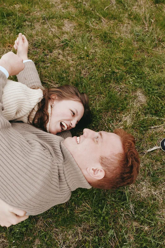 Smiling couple lying on grass, outdoor daylight, and cozy sweaters convey warmth, affection, and relaxed leisure, creating a candid lifestyle moment filled with happiness and connection on a natural,