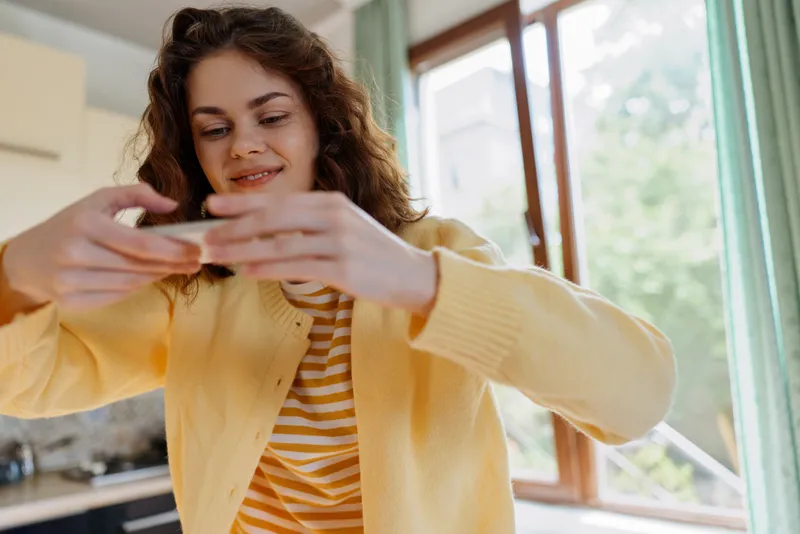 Woman in a yellow cardigan photographed at home, taking a casual selfie with a smartphone, bright natural light, cozy living space, cheerful mood, everyday lifestyle moment indoors.