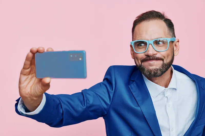 Professional man in a blue suit takes a confident selfie against a solid pink background, expressing upbeat mood, focus, and pride in a modern business setting.