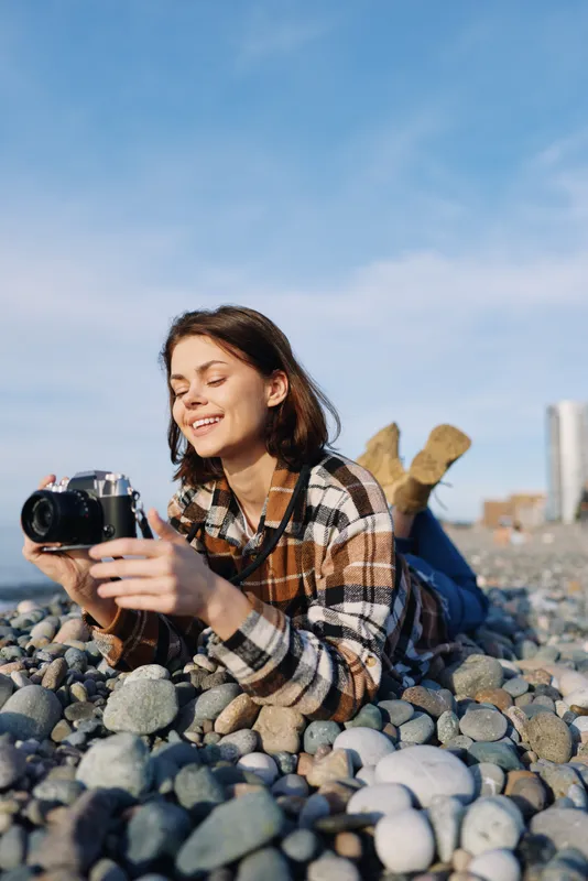 A woman photographer lies on a pebble beach, camera in hand, smiling as she captures candid seaside moments under clear blue skies and bright sun with relaxed posture and natural lighting
