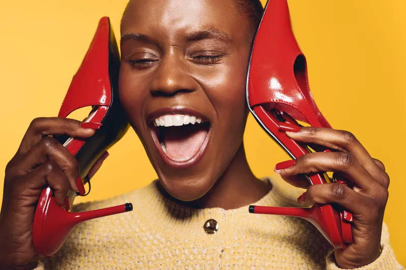 Happy African woman with dark skin holds red high heel shoes near her face, smiling with eyes closed on yellow background. Concept of fashion, joy, and beauty expression.