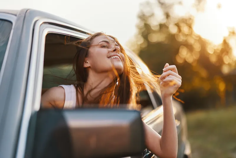 Young woman enjoying summer breeze with hair in the wind from car window, showcasing freedom and happiness during a scenic drive.
