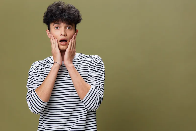 surprised man with curly hair wearing striped shirt, holding face with hands, expressive young adult portrait on plain olive background
