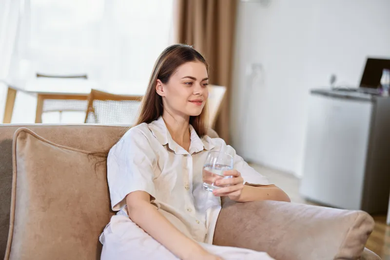 Woman on couch holding a glass of water at home, relaxed and sitting in the livingroom. Casual portrait of hydration and wellness, calm domestic lifestyle with soft natural light.