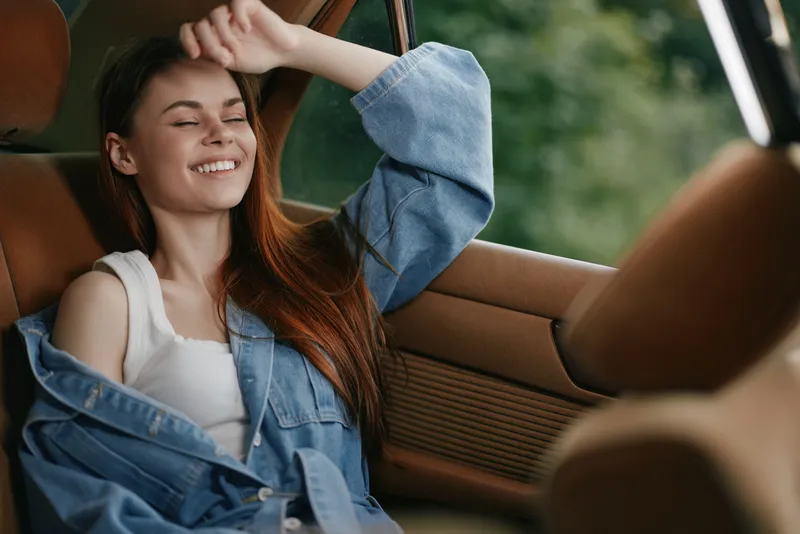 Young woman with long hair enjoying a sunny day in a car, wearing a denim jacket, relaxed and happy expression, surrounded by a natural green background.