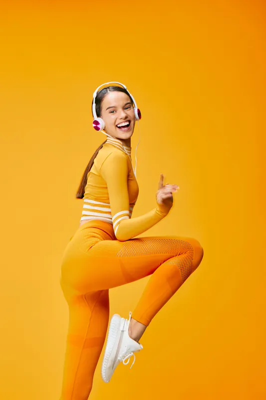 Female fitness enthusiast in bright orange sportswear with headphones, captured mid-jump against a vivid orange backdrop, showcasing energy, movement, and athletic style.