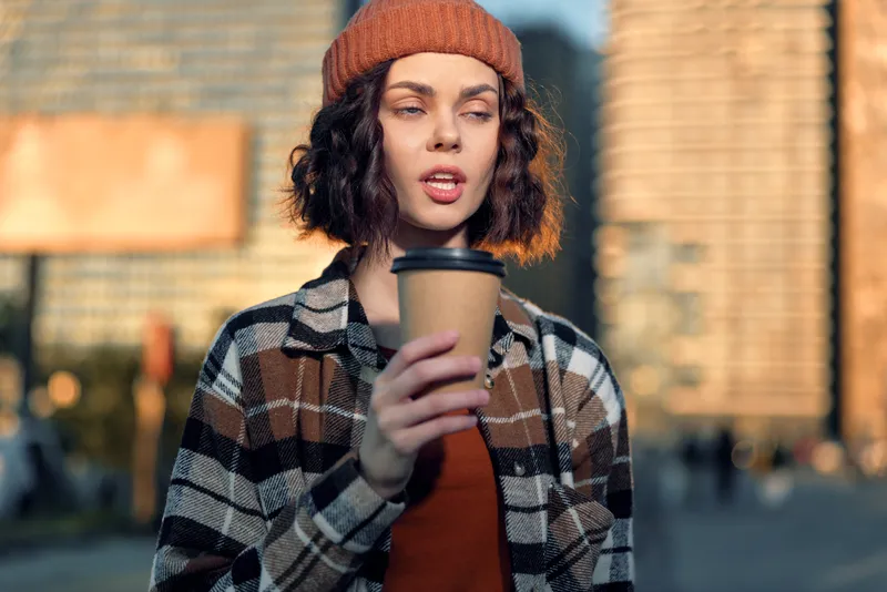 woman coffee urban portrait beanie candid authenticity golden captured in warm golden hour glow; thoughtful young adult holding takeaway cup in city, candid lifestyle and emotional storytelling.