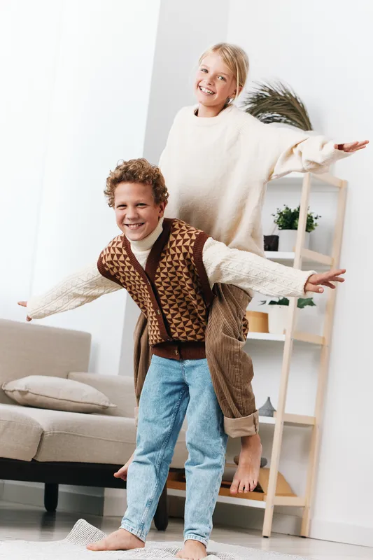 Two children joyfully dance indoors, arms wide, smiling in a bright living room with soft furnishings and a cozy, playful vibe.