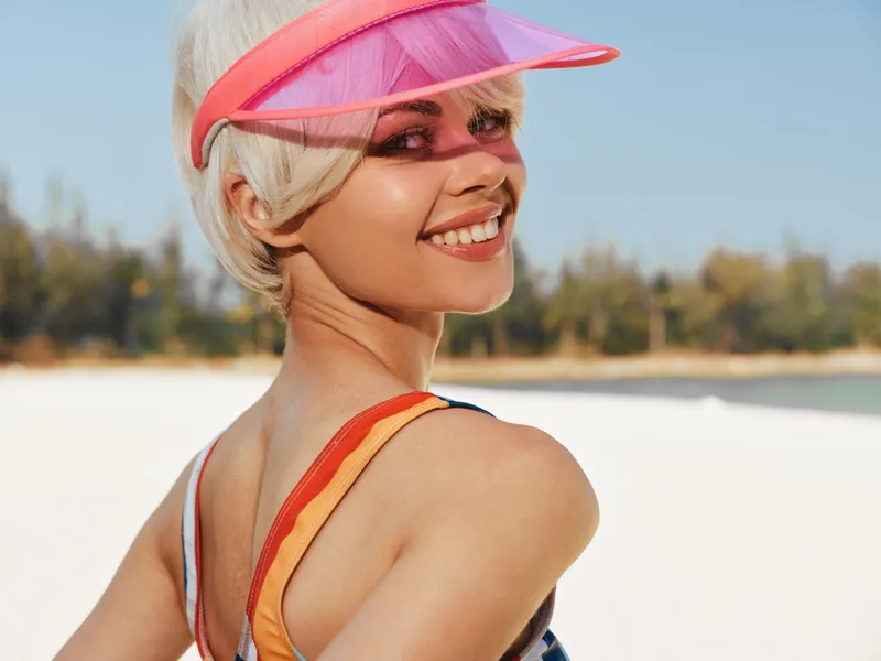 Smiling woman at sunny beach wearing a pink visor and colorful swimsuit, captured in bright natural light with sandy shore and blue sky, conveying carefree summer vibes and youthful energy.