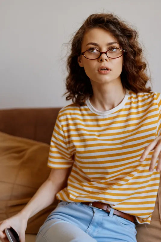 Young woman with curly brown hair and glasses sits on a soft sofa wearing a yellow striped tee, looking thoughtful in a warm, casual home setting.