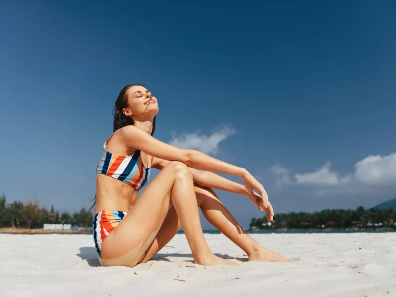 Sunlit beach scene featuring a woman in a striped bikini seated on white sand, eyes closed, basking in warm sunlight beneath a clear blue sky.