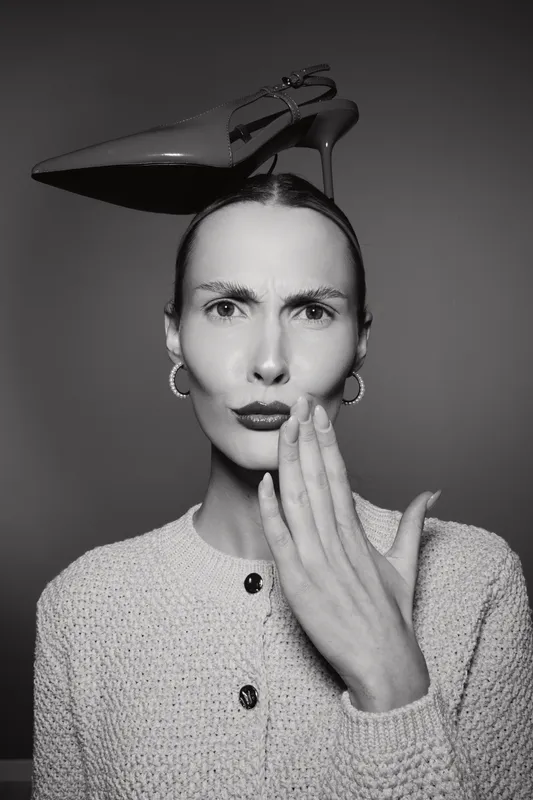 Portrait of slender Caucasian woman with light skin and slicked hair holding red high heel on her head, wearing earrings and knitted cardigan. Concept of fashion, style, and expression.